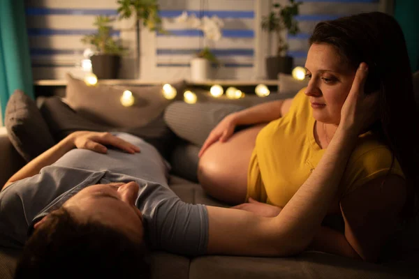 Una pareja casada yace en la cama y el marido le pega la mejilla a su esposa y se miran enamorados. Mujer en embarazo avanzado. — Foto de Stock