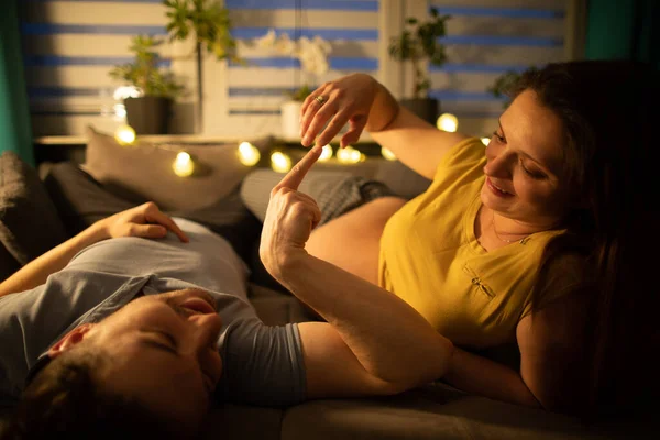 Una pareja casada se acuesta en una cama en un dormitorio y se tocan con sus dedos índice. La mujer tiene un embarazo avanzado.. — Foto de Stock