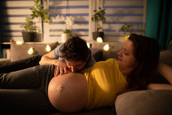 In the evening, lying on the bed, the husband kisses the belly of his pregnant wife. A woman in advanced pregnancy. — Stock Photo, Image