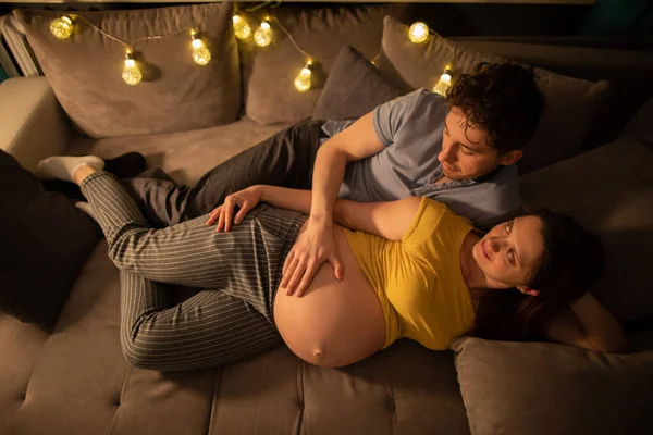 Ein Ehepaar liegt auf einem Bett im Schlafzimmer und der Ehemann berührt sanft den Bauch seiner schwangeren Frau. Eine Frau in fortgeschrittener Schwangerschaft. — Stockfoto