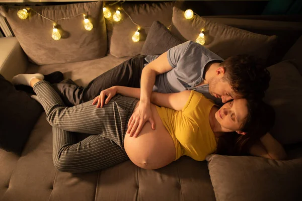 Una pareja casada está acostada en una cama en un dormitorio y el marido está tocando suavemente el vientre de sus esposas embarazadas. Una mujer en embarazo avanzado. — Foto de Stock