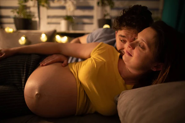 La esposa embarazada está acostada en la cama en el dormitorio y en el fondo se puede ver marido curioso. Mujer en embarazo avanzado. — Foto de Stock