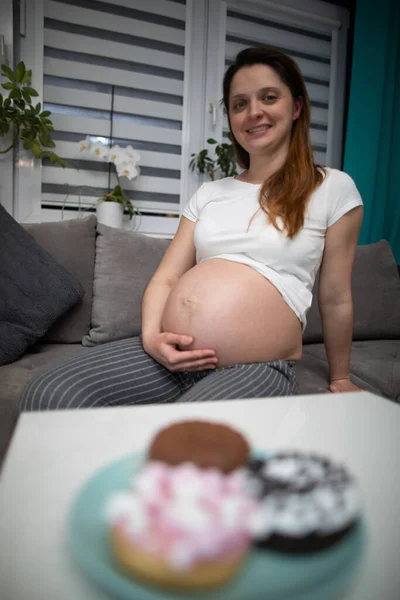 Una mujer embarazada se sienta en el sofá en casa y planea comer algunas rosquillas. Una mujer en embarazo avanzado. — Foto de Stock