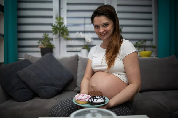 Una mujer embarazada se sienta en el sofá en casa y planea comer algunas rosquillas. Una mujer en embarazo avanzado. — Foto de Stock