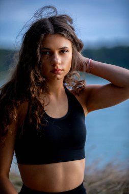 Genç bir kadın sakin bir gölün kenarında, atletik kıyafetlerini ve doğal güzelliğini göstererek rahatça poz veriyor.