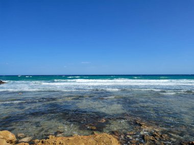 Nefes kesici bir plaj sahnesi berrak mavi suları, yumuşak beyaz dalgaları ve yukarıda parlak güneşli gökyüzünü gözler önüne seriyor.