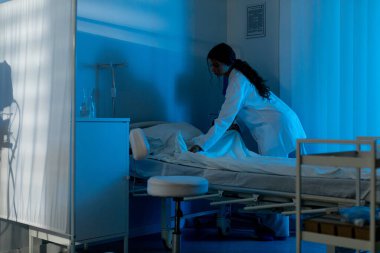 Bakıcı, hafif mavi parıltı altında konfor sağlar, hizmetli nazikçe yatağı monitör olarak düzenler ve tüp yumuşak bir ışık altında güven verici bir varlık sağlar