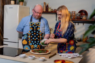 Beyaz çift mutfakta yemek hazırlıyor, seramik tabaklar üzerinde taze otlar düzenliyor, ahşap tezgah, sıcak ışıklandırma, günlük ev yemeği havası.