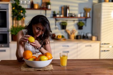 Mutfakta turuncu kaseyi düzenleyen kadın portakal suyu seçen eller, kâsenin yanında bir bardak meyve suyu, ahşap masa üstü, güneş ışığı çizgileri, nazik konsantrasyon, günlük kıyafet.,