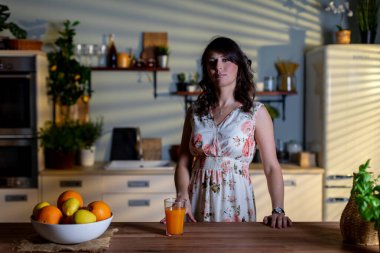 Mutfakta portakallı beyaz bir kadın, ahşap tezgahın arkasında elinde turunçgil ve bir bardak meyve suyu, yumuşak pencere ışığı çizgili gölgeler yaratıyor.,