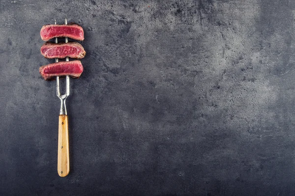 Skivor av nötkött ryggbiff på kött gaffel på konkreta bakgrund — Stockfoto