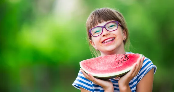 Barn äter vattenmelon. Barnen äter frukt i trädgården. Pre Teen flicka i trädgården som håller en bit vatten melon. lycklig flicka unge äter vattenmelon. Flicka unge med gaser och tänder hängslen — Stockfoto