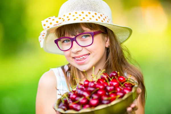 Criança com cerejas. Menina com cerejas frescas. Jovem menina loira caucasiana bonito vestindo dentes aparelhos e óculos. Retrato de uma jovem sorridente com tigela cheia de cerejas frescas — Fotografia de Stock