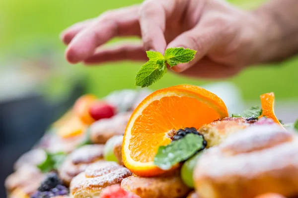 Kocken förbereder en platta av kakor med färsk frukt. Han arbetar på ört dekoration. Utomhus trädgårdsfest — Stockfoto