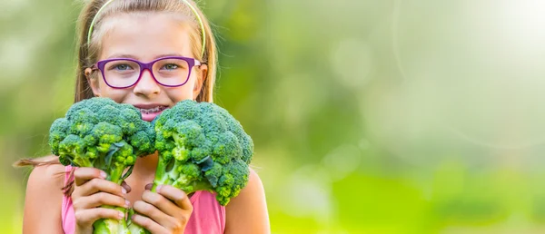 Chica linda divertida sosteniendo en las manos la col roja y el brócoli. Fondo borroso en el jardín. Pre-adolescente joven con gafas y aparatos ortopédicos — Foto de Stock