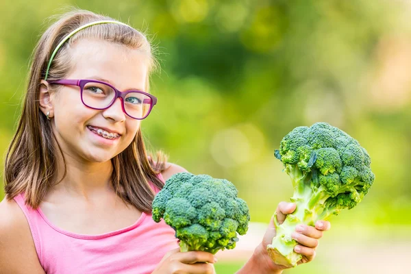 Chica linda divertida sosteniendo en las manos la col roja y el brócoli. Fondo borroso en el jardín. Pre-adolescente joven con gafas y aparatos ortopédicos — Foto de Stock