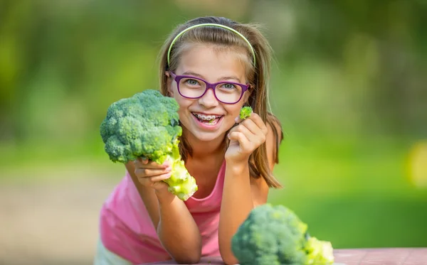 Chica linda divertida sosteniendo en las manos la col roja y el brócoli. Fondo borroso en el jardín. Pre-adolescente joven con gafas y aparatos ortopédicos — Foto de Stock