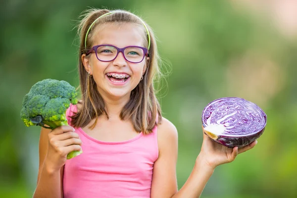 Chica linda divertida sosteniendo en las manos la col roja y el brócoli. Fondo borroso en el jardín. Pre-adolescente joven con gafas y aparatos ortopédicos — Foto de Stock