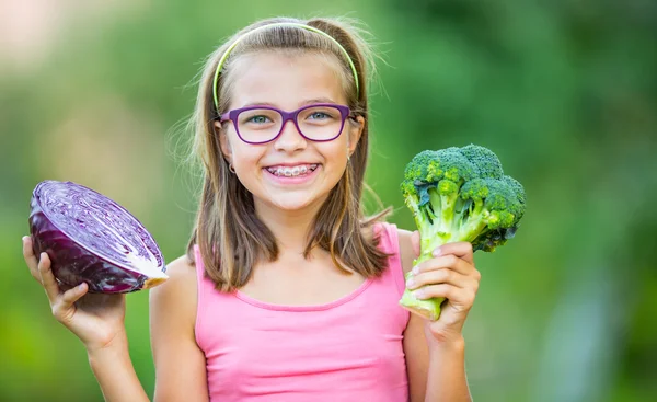 Chica linda divertida sosteniendo en las manos la col roja y el brócoli. Fondo borroso en el jardín. Pre-adolescente joven con gafas y aparatos ortopédicos — Foto de Stock