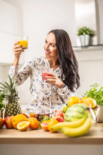 Good-looking brunette looks at a glass of yellow juice in one hand, holding red juice in the other hand.