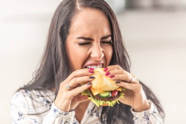 Büyük bir hamburgeri ısırmak için mümkün olduğunca çok ağzını açmaya çalışan bir kız..
