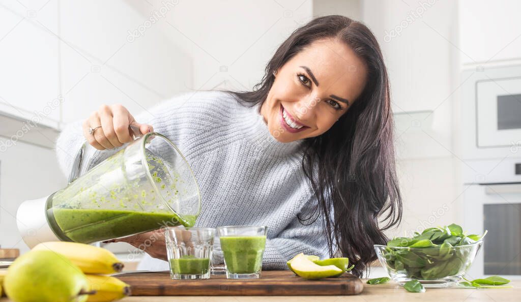 Similing female pours freshly made green smoothie into glasses in the kitchen.