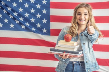 Mutlu üniversite ya da üniversite öğrencisi çalışma materyalleri ve arkasında Amerikan bayrağı olan bir elma taşıyor..