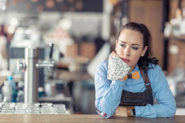 Sad female business owner holds face mask in her hand and leans against the bar in her empty cafe.