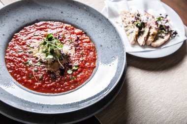 Kırmızı domates çorbası yeşil ve peynirli bir tabakta servis ediliyor. Yanında da ekmek dilimleri..