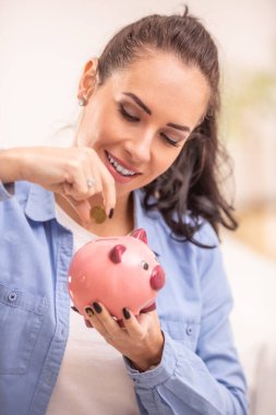 Elinde bakır para tutan esmer bir kadın tarafından pembe bir kumbaraya para yatırılıyor..