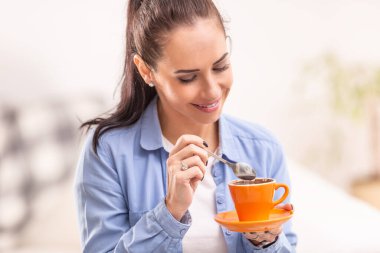 Genç bir kadın sabah kahvesini elinde bir kaşıkla turuncu bir fincanda tutuyor..