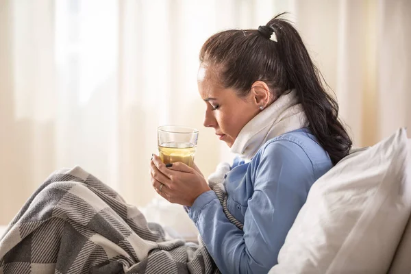 Woman shivering from cold heats herself by holding a cup of hot tea in her hands on a sofa in the house.