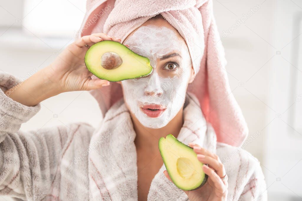 Woman ina dressing gown and towel on her hair wears white face mask and halved avocado over her eye.