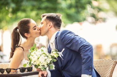 Joyful couple sitting and kissing each other after wedding. Bride holds elegant bouquet.