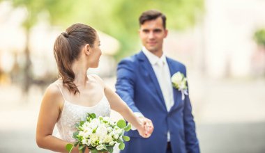 Attractive bride is holding her husbands hand while looking at him and in the other hand she holds lovely bouquet. They wear wedding robes.