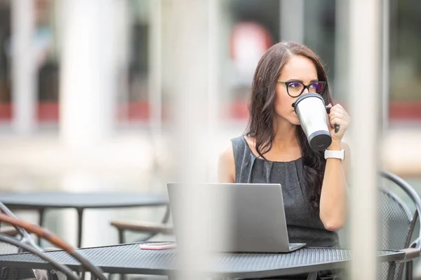 Beautiful brunette sipping coffee from sustainable coffee mug outdoors while she works on the laptop in an offce enviroment.