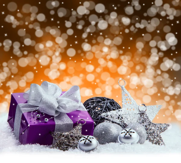 Μωβ πακέτο Χριστουγέννων, δώρο της μια ασημένια κορδέλα. Τρίγωνα κάλαντα, τα ασημένια Χριστούγεννα μπάλες και Χριστούγεννα αστέρια θέσει στο χιόνι. Αφηρημένη πολύχρωμα θολή φόντο χώρο. — Φωτογραφία Αρχείου
