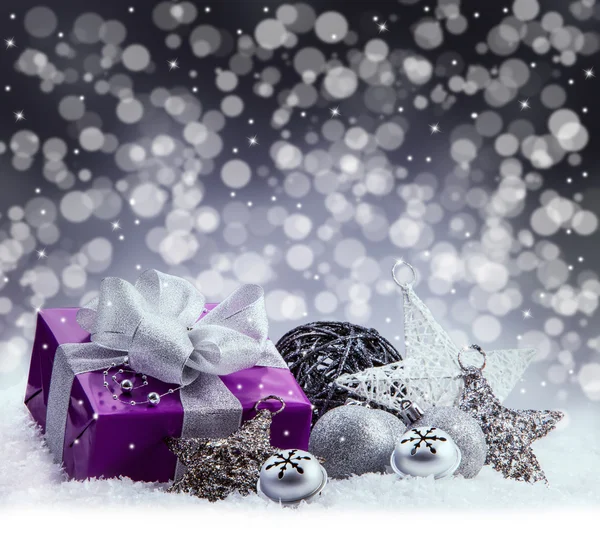 Purple Christmas package , gift of a silver ribbon. Jingle bells , silver christmas balls and christmas stars put on snow. Abstract multicolored blurred background space.