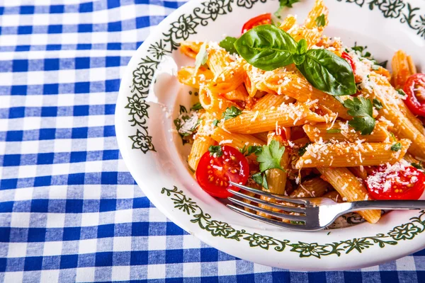 Prato com caneta de macarrão Molho de bolonhesa tomates cereja salsa superior e folhas de manjericão em xadrez toalha de mesa azul . — Fotografia de Stock