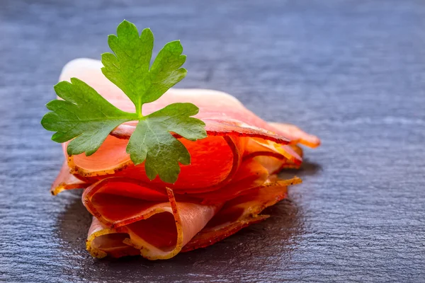 Gekrulde segmenten van Delicious Prosciutto met peterselie bladeren op graniet bord. — Stockfoto