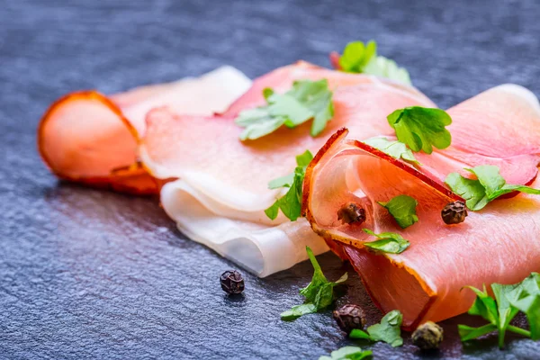 Gekrulde segmenten van Delicious Prosciutto met peterselie bladeren op graniet bord. — Stockfoto