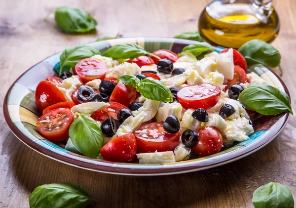 Caprese. Caprese salad. Italian salad. Mediterranean salad. Italian cuisine. Mediterranean cuisine. Tomato mozzarella basil leaves black olives and olive oil on wooden table. — Stock fotografie