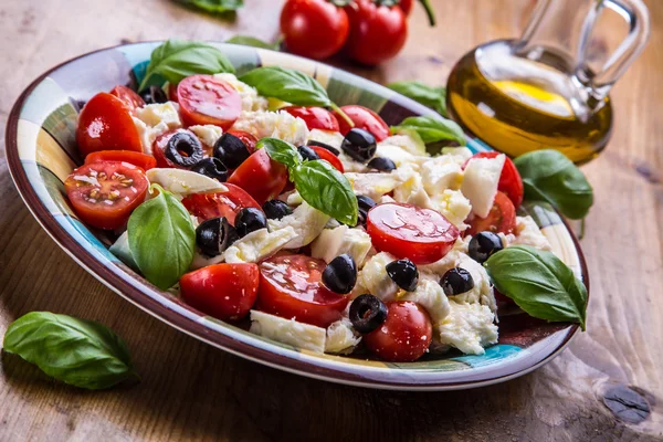 Caprese. Caprese salad. Italian salad. Mediterranean salad. Italian cuisine. Mediterranean cuisine. Tomato mozzarella basil leaves black olives and olive oil on wooden table. — Stock Fotó