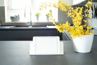 Modern mutfağın manzarası. Mutfak masasının üzerinde beyaz boş ekran olan tabletin bir kopyası. Uzayı kopyala Forsythia 'nın ön plandaki beyaz bir vazoda, arka plandaki güneş ışığıyla aydınlatılmış dallarda. İtalyan iç tasarımı