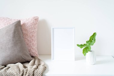 Beyaz masaya boş beyaz çerçeve modeli. Yumuşak renkli yastıklar, ekoseli vazo ve arum italikum yapraklı beyaz vazo. Modern ve asgari yaşam tarzı. İskandinav iç tasarımı