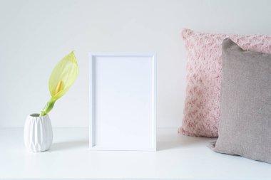 Beyaz masaya boş beyaz çerçeve modeli. Yumuşak renkli yastıklar ve arum italikum çiçekli beyaz vazo. Modern ve asgari yaşam tarzı. İskandinav iç tasarımı