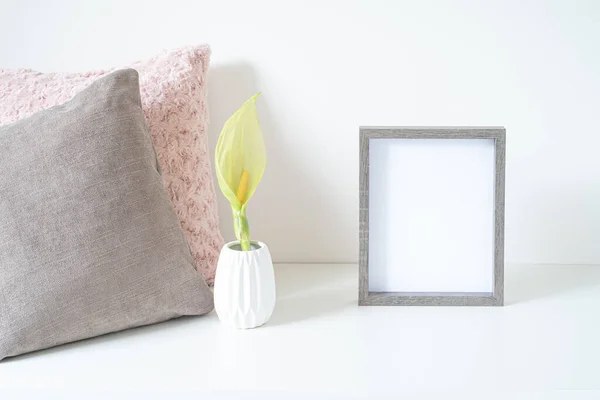 Beyaz masaya boş ahşap çerçeve modeli. Yumuşak renkli yastıklar ve arum italikum çiçekli beyaz vazo. Modern ve asgari yaşam tarzı. İskandinav iç tasarımı