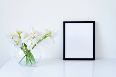 Boş siyah çerçeve modeli. Beyaz süsen çiçekleri kürenin içinde beyaz masanın üzerinde cam vazo şeklinde. Beyaz arkaplan, minimal ve zarif boşluk