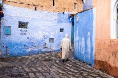 Chefchaouen, Fas - 26 Mart 2024: Bulutlu bir günde, Fas 'ın mavi şehri olan Chefchaouen' in sokaklarında tanınmayan geleneksel giysiler içinde yürüyen kişi
