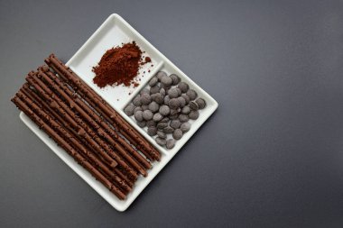Uzun çikolata çubukları, çikolata düğmeleri ve gri arka planda kare bir tabakta kakao tozu..